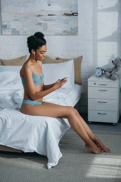 Африканская американка в нижнем белье слушает музыку с наушниками и смартфоном в спальне — стоковое фото