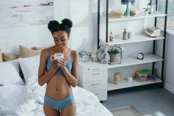 Schöne lächelnde afrikanisch-amerikanische Frau in Unterwäsche, die morgens mit einer Tasse Kaffee im Schlafzimmer steht — Stockfoto