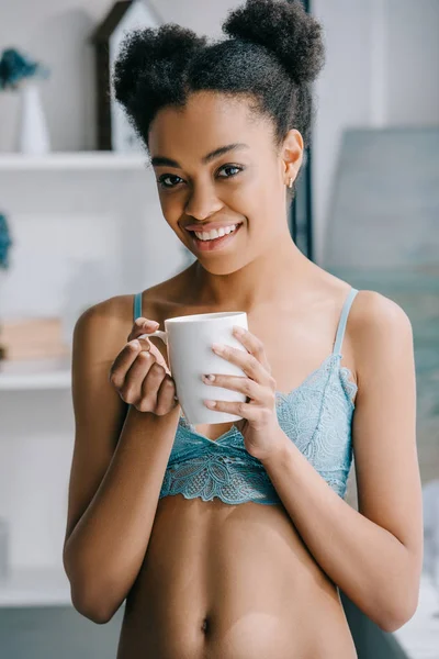 Hermosa sonriente afroamericana chica en lencería celebración taza de té en la mañana - foto de stock