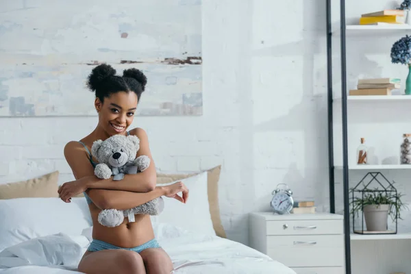 Улыбающаяся африканская американка, обнимающая плюшевого мишку, сидя утром на кровати — стоковое фото