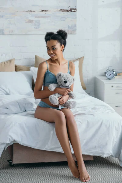 Счастливая африканская американка в нижнем белье держит плюшевого мишку в спальне утром — стоковое фото
