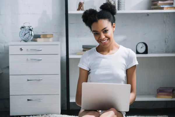 Chica afroamericana feliz usando el ordenador portátil blanco sentado en el piso en el dormitorio - foto de stock