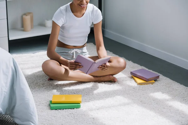 Vista recortada del estudiante afroamericano leyendo libro en el piso en el dormitorio - foto de stock