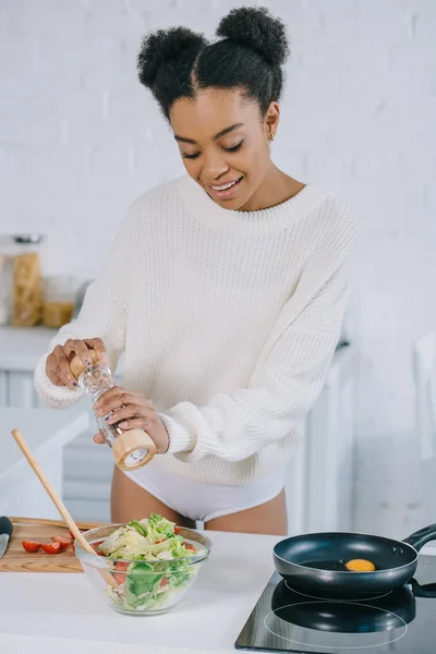 Beautiful young woman with pepper mill peppering salad at kitchen — Stock Photo