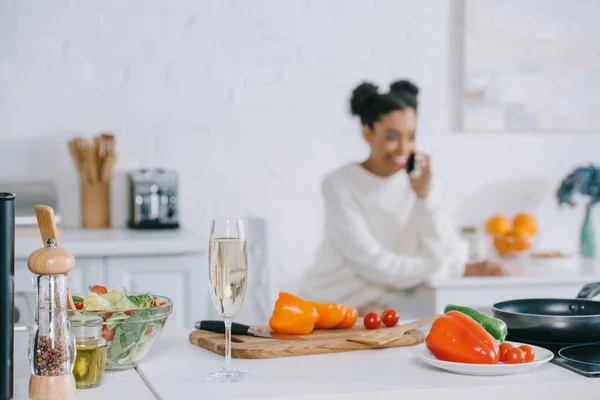 Jeune femme souriante floue parlant par téléphone avec des légumes et une coupe de champagne au premier plan à la maison — Photo de stock