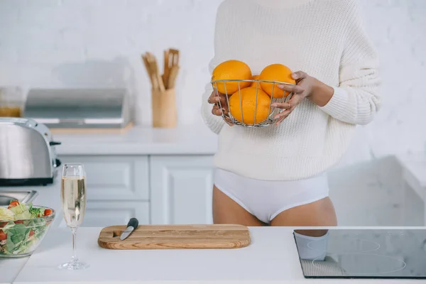 Tiro recortado de mujer en ropa interior y suéter con cesta de naranjas frescas en la cocina - foto de stock