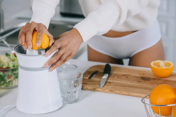 Tiro recortado de la mujer en ropa interior y suéter exprimiendo jugo de naranja fresca en la cocina - foto de stock