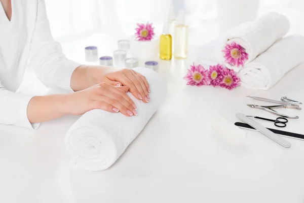 Partial view of woman holding hands at table with towels, flowers, candles, aroma oil bottles and instruments for manicure in beauty salon — Stock Photo