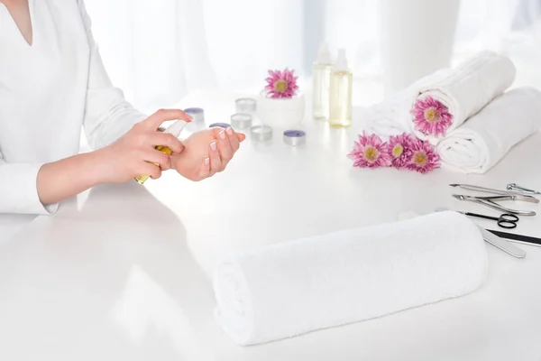 Imagem cortada da mulher usando óleo de aroma à mesa com toalhas, flores, velas e instrumentos para manicure no salão de beleza — Fotografia de Stock