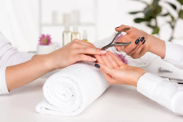 Image recadrée de la femme recevant manucure par esthéticienne avec coupe-ongles à table avec des fleurs et des serviettes dans le salon de beauté — Photo de stock