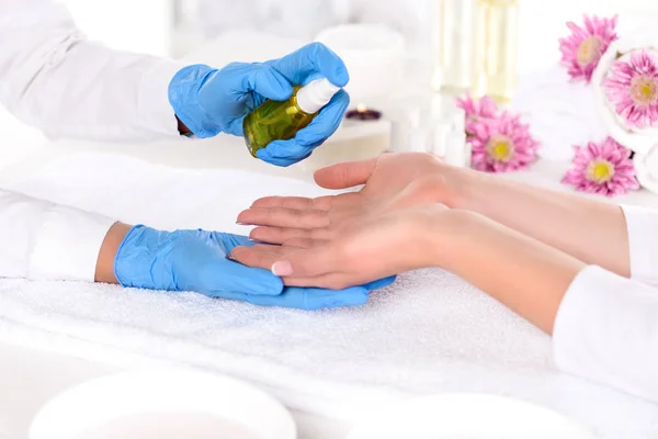 Cropped image of manicurist in latex gloves spraying aroma oil on hands of woman in beauty salon — Stock Photo
