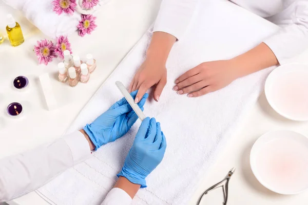 Top view of beautician in latex gloves doing manicure to woman by nail file at table with flowers, towels, nail clippers, candles, nail polishes and aroma oil bottles — Stock Photo