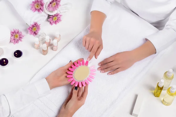 Top view of manicurist holding samples of nail varnishes while woman pointing by finger at table with aroma oil bottles, candles, towels, flowers and nail polishes in beauty salon — Stock Photo