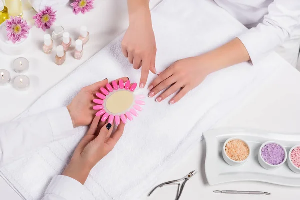 Cropped image of manicurist holding samples of nail varnishes while woman pointing by finger at table with aroma oil bottles, candles, towels, flowers, nail polishes, sea salt and nail clippers — Stock Photo