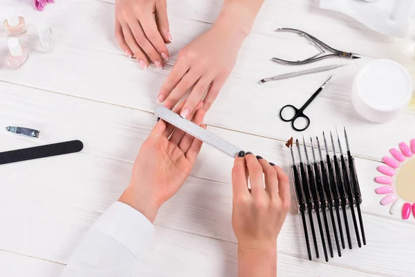 Обрізане зображення косметолога, який робить манікюр за допомогою нігтів для жінки за столом з кремом, лаками для нігтів, ножицями, кутикулою, ножицями для нігтів, зразками лаків для нігтів — стокове фото