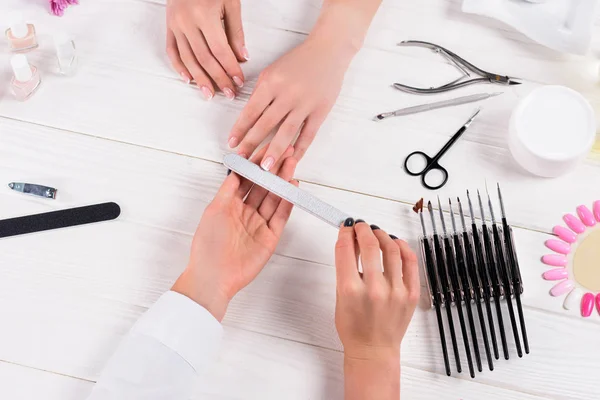 Image recadrée de l'esthéticienne faisant manucure par lime à ongles à la femme à table avec crème, vernis à ongles, ciseaux, poussoir à cuticules, coupe-ongles, échantillons de vernis à ongles — Photo de stock