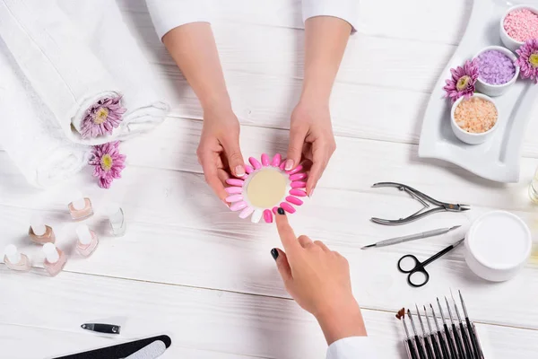 Manicurista mostrando muestras de esmaltes de uñas a la mujer en la mesa con flores, toallas, esmaltes de uñas, limas de uñas, cortauñas, sal marina, crema, empujador de cutículas y tijeras - foto de stock