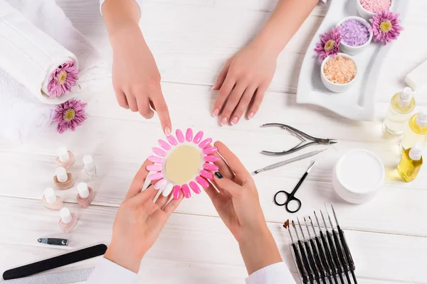 Vue du dessus de la femme pointant sur des échantillons de vernis à ongles dans les mains de manucure à table avec vernis à ongles, limes à ongles, coupe-ongles, poussoir à cuticules, sel de mer, fleurs, bouteilles d'huile aromatique et échantillons de vernis à ongles — Photo de stock