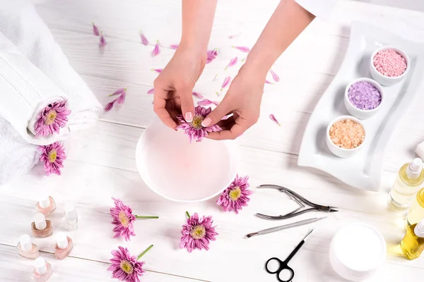 Partial view of woman throwing petals in bath for nails at table with flowers, towels, colorful sea salt, aroma oil bottles, nail polishes, cream container and tools for manicure in beauty salon — Stock Photo