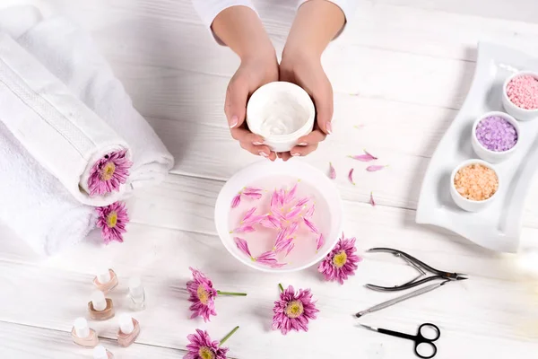 Tiro recortado de la mujer que sostiene la crema sobre la mesa con baño para las uñas, flores, toallas, sal marina colorida, esmaltes de uñas y herramientas para la manicura en el salón de belleza - foto de stock