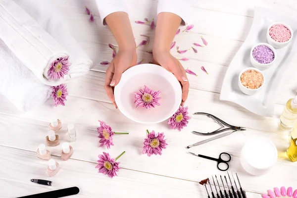 Teilbild einer Frau, die ein Nagelbad über dem Tisch mit Blumen, Handtüchern, buntem Meersalz, Aromaölflaschen, Nagellacken, Cremebehältern und Manikürwerkzeugen im Schönheitssalon hält — Stockfoto