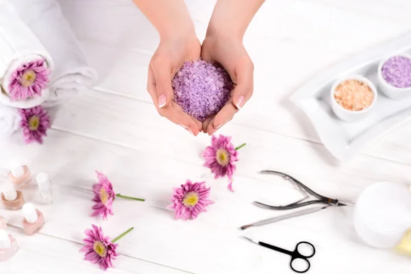 Teilansicht einer Frau, die Meersalz mit Blumen, Handtüchern, Nagellacken, Cremebehältern und Manikürwerkzeugen im Schönheitssalon über den Tisch hält — Stockfoto