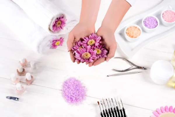 Imagen recortada de la mujer sosteniendo flores sobre la mesa con toallas, esmaltes de uñas, sal marina de colores, recipiente de crema y herramientas para la manicura en el salón de belleza - foto de stock
