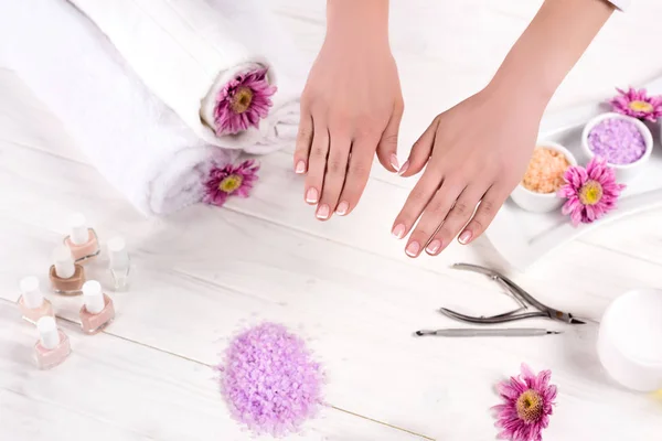 Visão parcial das mãos femininas à mesa com toalhas, flores, esmaltes, sal marinho colorido, recipiente de creme e ferramentas para manicure no salão de beleza — Fotografia de Stock