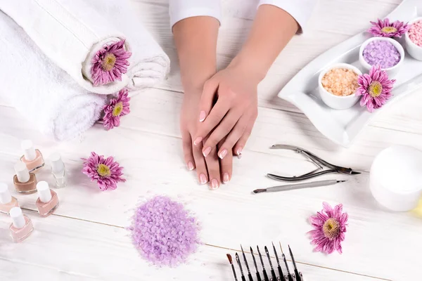 Tiro recortado de mãos femininas à mesa com toalhas, flores, esmaltes, sal marinho colorido, recipiente de creme e ferramentas para manicure no salão de beleza — Fotografia de Stock