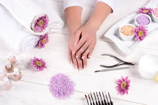 Plan recadré de mains féminines à table avec serviettes, fleurs, vernis à ongles, sel de mer coloré, récipient à crème et outils pour la manucure dans le salon de beauté — Photo de stock