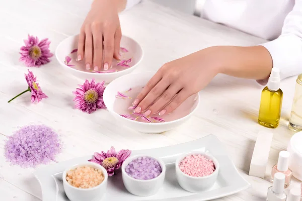 Vista parziale della donna che riceve il bagno per le unghie a tavola con fiori, sale marino colorato, contenitore di crema, bottiglie di olio aromatico e smalti per unghie nel salone di bellezza — Foto stock