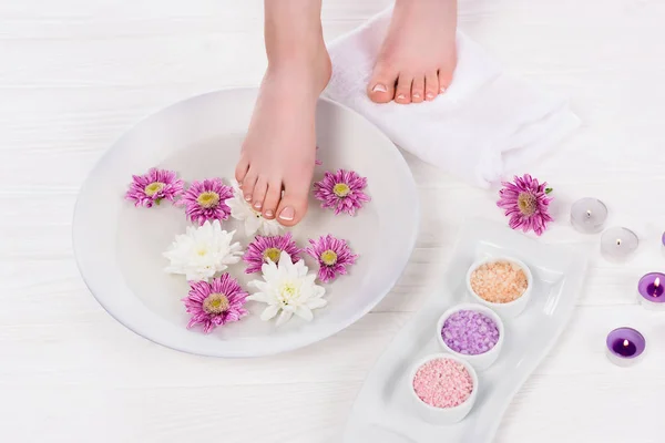 Tiro cortado de mulher descalça recebendo banho para unhas com sal marinho colorido e flores perto de velas de aroma no salão de beleza — Fotografia de Stock