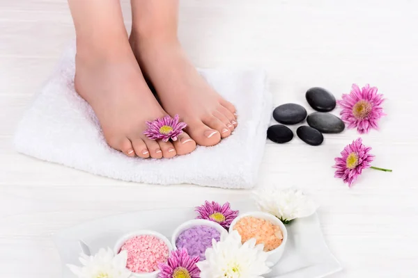 Image recadrée de femme pieds nus sur le traitement spa avec serviette, fleurs, sel de mer coloré et pierres de spa — Photo de stock