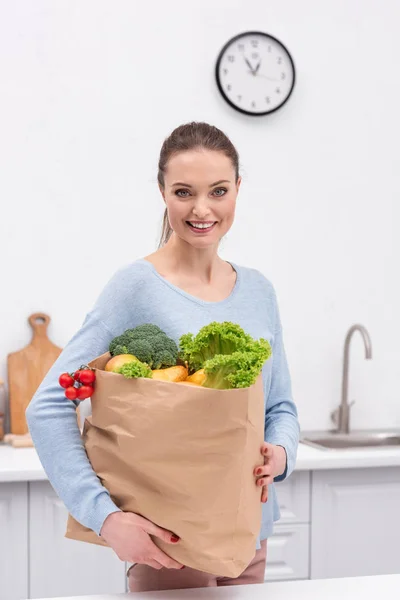Счастливая взрослая женщина с бумажным пакетом с овощами и фруктами на кухне — стоковое фото
