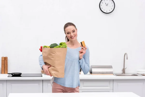 Happy adult woman with paper bag and credit card at kitchen — Stock Photo