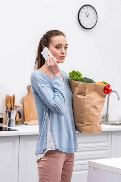 Beautiful adult woman with paper bag talking by phone at kitchen — Stock Photo