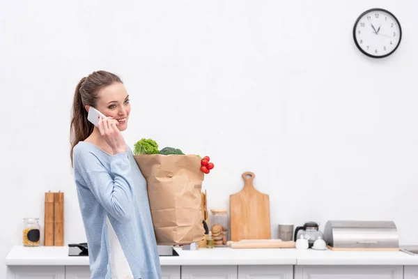 Happy adult woman with paper bag talking by phone at kitchen — Stock Photo
