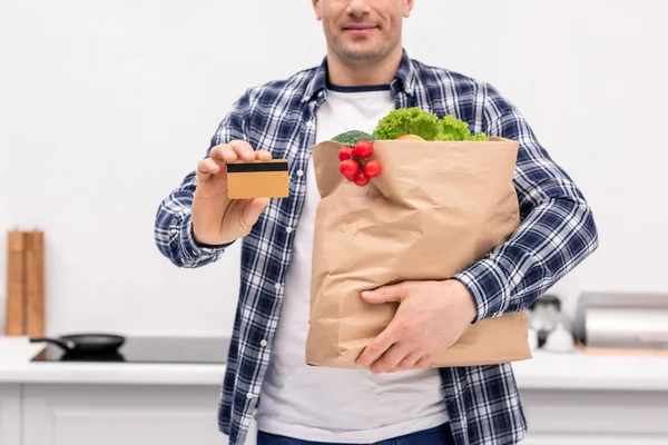 Обрезанный снимок улыбающегося взрослого мужчины с сумкой из продуктового магазина и кредиткой на кухне — стоковое фото