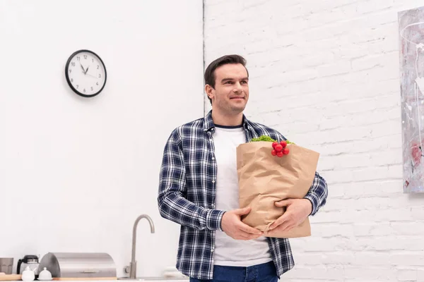 Sonriente hombre adulto llevando bolsa de la tienda de comestibles en la cocina - foto de stock
