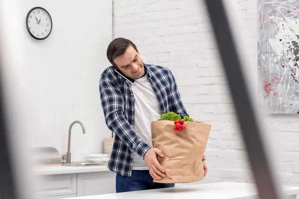 Lächelnder erwachsener Mann trägt Einkaufstasche und telefoniert in der Küche — Stockfoto