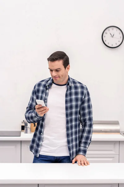 Усміхнений дорослий чоловік використовує смартфон на кухні — стокове фото
