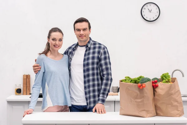 Feliz pareja adulta con bolsas de papel de la tienda de comestibles abrazando en la cocina y mirando a la cámara - foto de stock