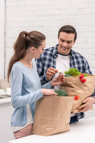 Улыбающаяся взрослая пара с бумажными пакетами из продуктового магазина на кухне — стоковое фото