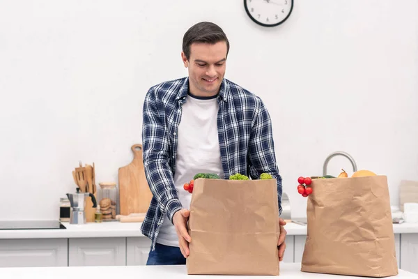 Hombre adulto feliz con bolsas de papel de supermercado en la cocina - foto de stock