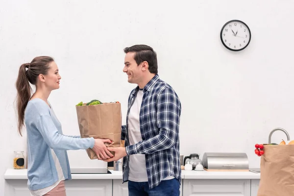 Happy husband taking paper bag with vegetables from wife at kitchen — Stock Photo