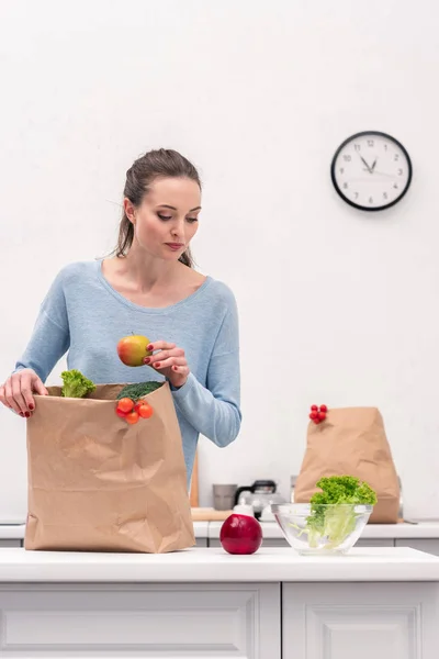 Adult woman taking fruits and vegetables out of paper bag at kitchen — Stock Photo