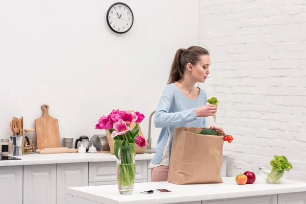 Beautiful adult woman taking fruits and vegetables out of paper bag at kitchen — Stock Photo