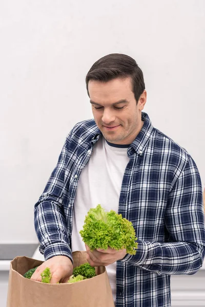 Smiling adult man unpacking grocery store paper bag at kitchen — Stock Photo
