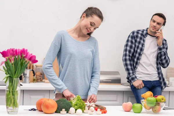 Happy adult man talking by phone and looking at his wife while she preparing dinner — Stock Photo