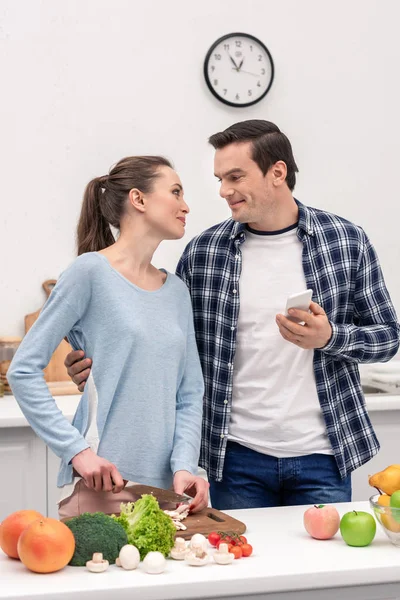 Homem adulto feliz com smartphone abraçando sua esposa enquanto ela prepara o jantar — Fotografia de Stock